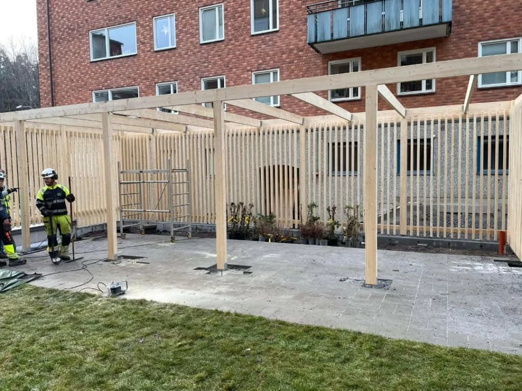 Stor ljus träpergola med ribbvägg under byggnation på stenlagd uteplats framför tegelhus, arbetare i skyddsutrustning